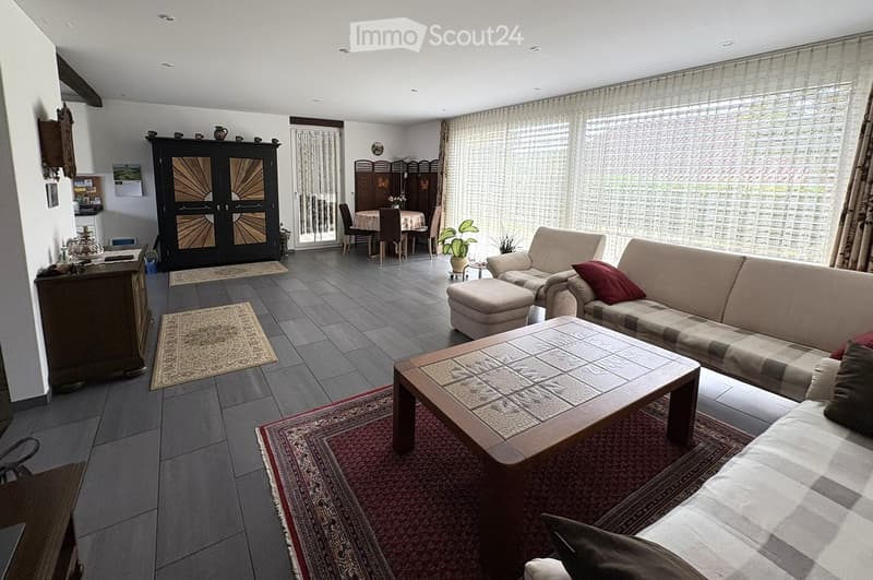 Bright living room with large windows, grey sofa, coffee table, ottoman, dining area, and decorative cabinet.