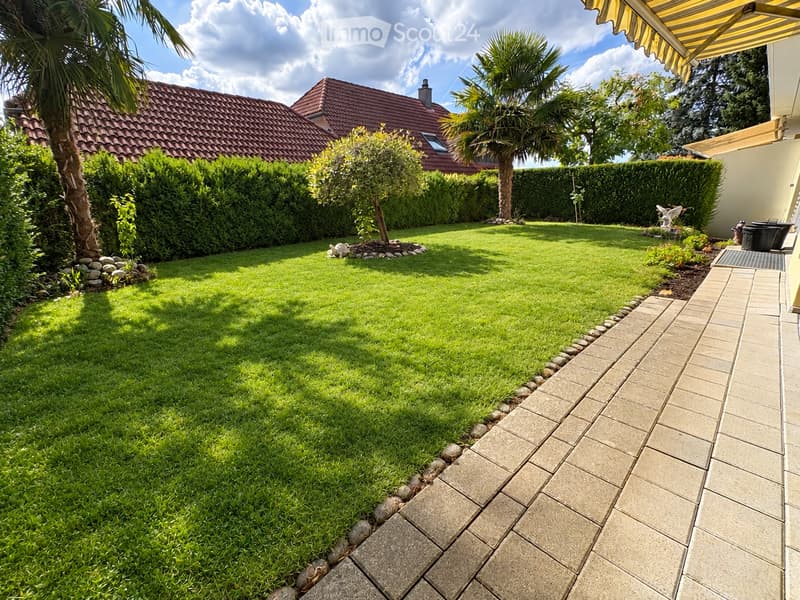 Well-maintained garden with a neatly trimmed lawn, a paved pathway, and surrounding greenery including palm trees and shrubs. A small decorative fountain is visible near the center.