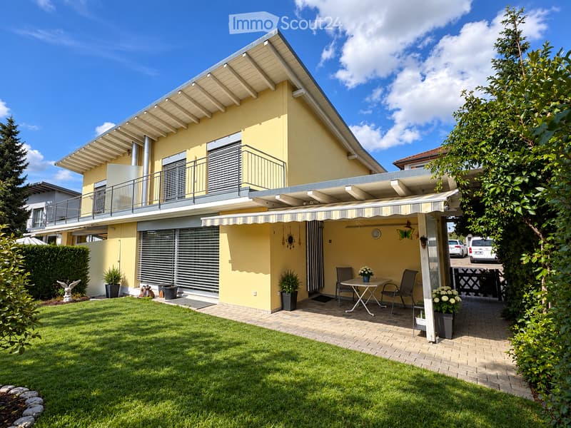 A modern two-story house with a yellow facade, featuring a covered patio area with a table and chairs, surrounded by a well-maintained lawn and garden.