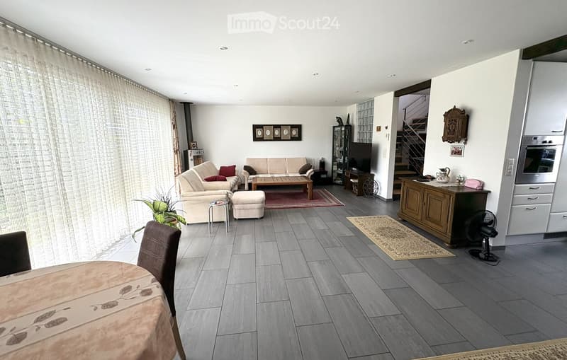 Open-plan living and dining area with grey tiled flooring, large windows with curtains, a sofa set, coffee table, ottoman, dining table with chairs, a fan, and an oven in the background.