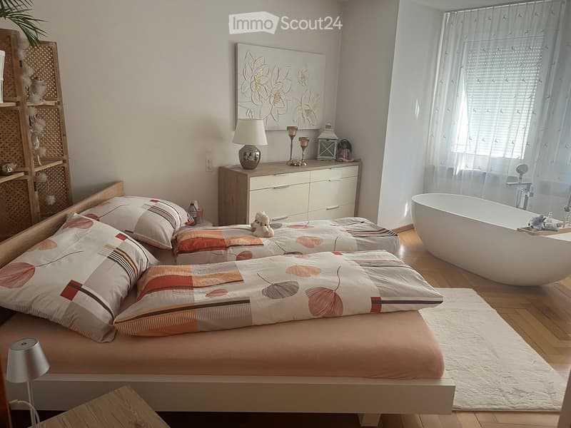 A modern bedroom with a large bed, nightstand, lamp, and a bathtub visible through a glass partition. The room features a wooden floor, white walls, and decorative items on the nightstand.