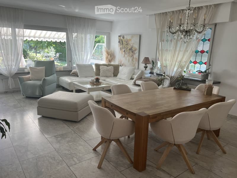 Bright living and dining area with large windows, white walls, a chandelier, a sofa, ottoman, coffee table, dining table with chairs, and decorative items.