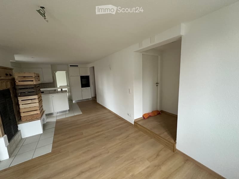 Empty hallway with wooden flooring, white walls, and a visible kitchen area in the background. There is a stack of wooden crates and a fireplace on the left side.