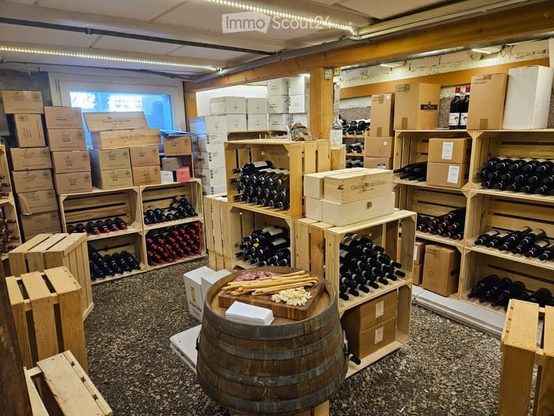 Wine cellar with wooden shelves holding rows of wine bottles, wooden crates, and barrels. A large wooden barrel serves as a table with cheese and glasses.