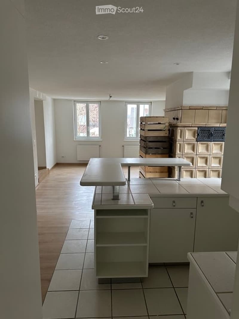 A modern kitchen with white cabinets, a central island, and a tiled floor. The space is well-lit with natural light from large windows, and includes a simple table and shelves.
