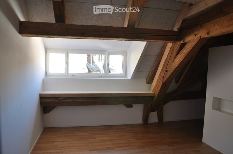 Empty attic room with wooden beams, white walls, wooden floor, and a window offering a view of the outside.