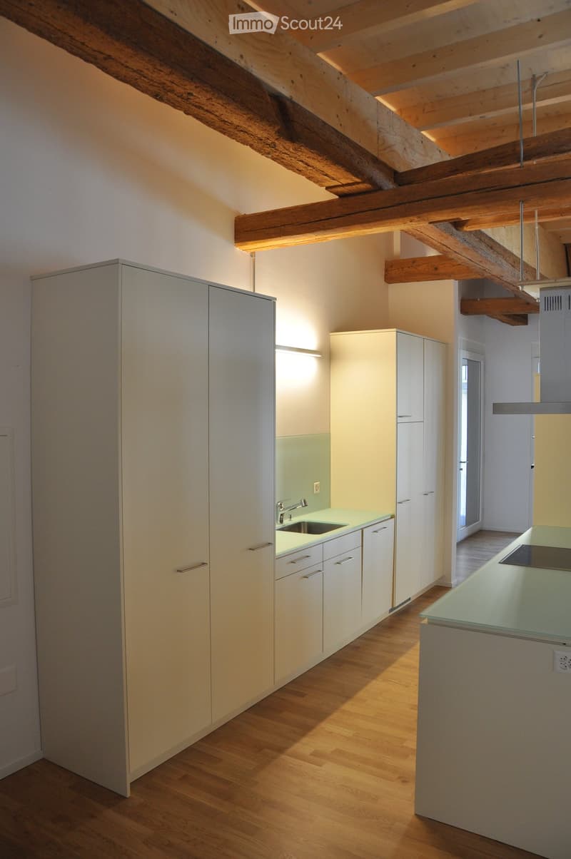 Modern kitchen with light-colored cabinets, wooden flooring, and a green countertop. Features exposed wooden beams on the ceiling and built-in storage.