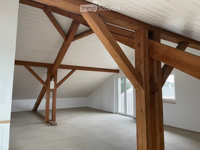 An empty room with exposed wooden beams and a slanted ceiling, featuring white walls, a tiled floor, and large windows with blinds.