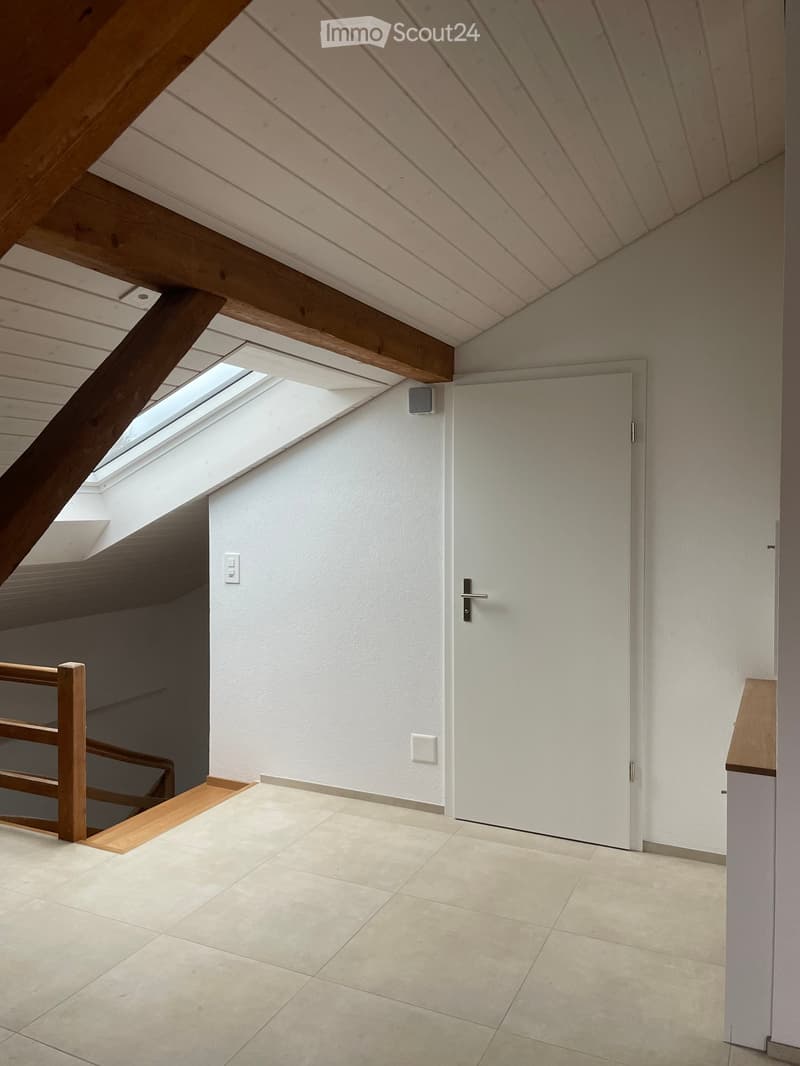 Bright hallway with wooden beams, tiled floor, white walls, and a skylight window at the top, leading to a closed white door.