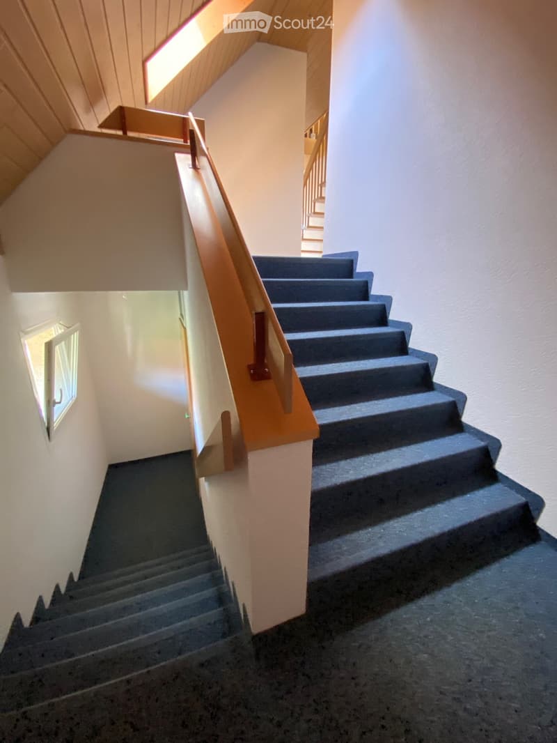 Staircase with wooden handrail and blue carpeted steps, leading to an upper level with visible lighting and walls.