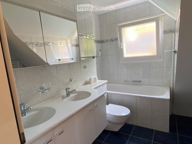 Modern bathroom with double sink, toilet, bathtub, and large mirror. Light grey tiles and dark blue floor.