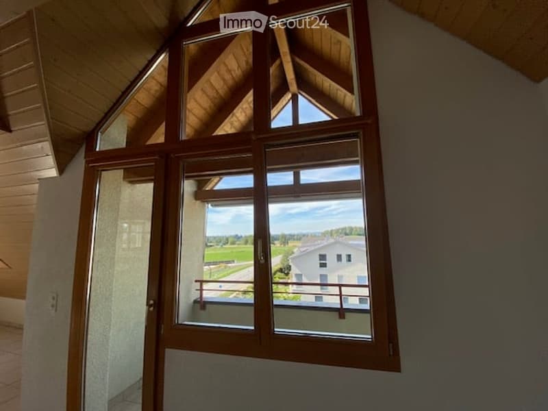 Bright hallway with wooden ceiling beams and large windows offering a scenic view of a green field and neighboring building.