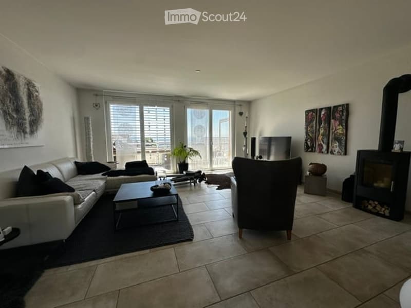 Modern living room with large windows and balcony access, featuring a white sectional sofa, dark coffee table, armchair, fireplace, TV, and a potted plant.