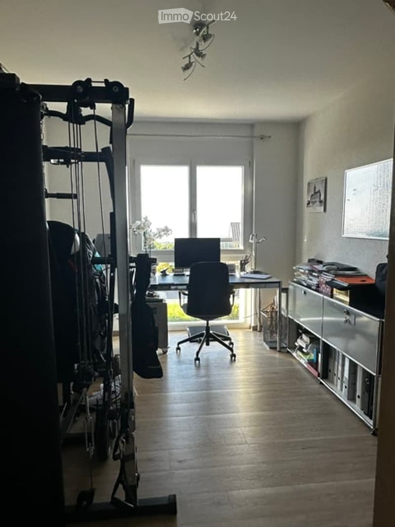 A modern, well-lit room with a wooden floor, a desk with a laptop and chair, a fitness machine on the left, and a cabinet with drawers on the right. Large windows allow natural light to fill the space.