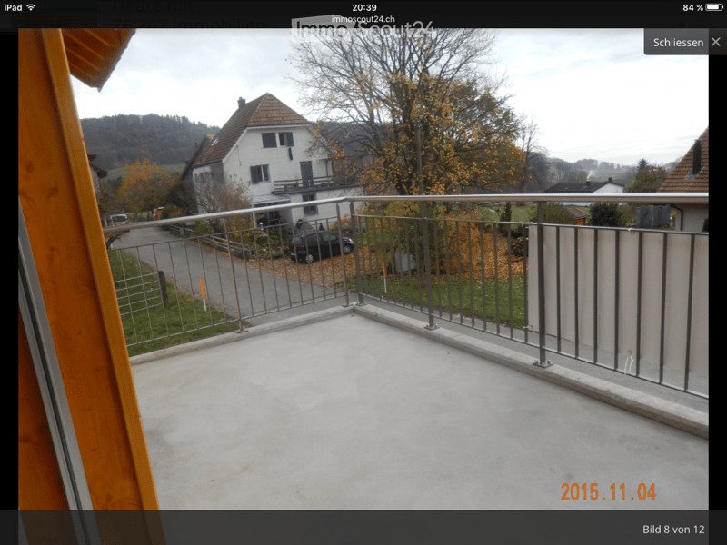 Balcony with metal railing, concrete floor, overlooking a residential area with houses, trees, and a parked car.