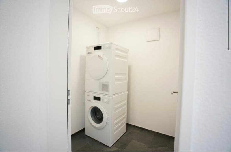 Compact room with a stacked washing machine and dryer unit against a white wall, featuring a dark tiled floor and a closed door.