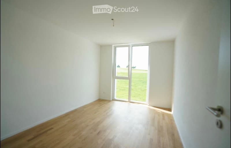 Empty room with light wooden flooring, white walls, and a large window with a view of a green field.