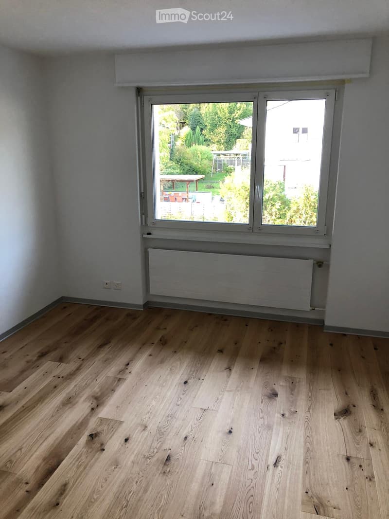 Empty room with light wooden flooring, white walls, a large window with a view of a garden, and a radiator below the window.