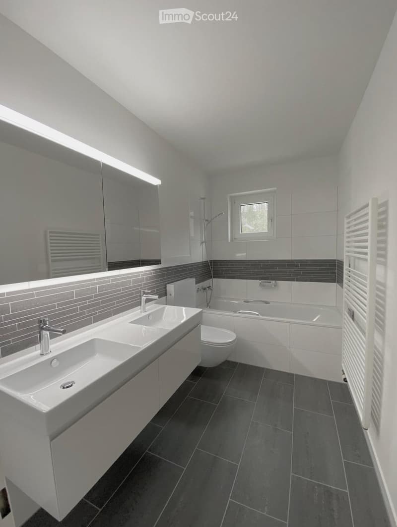 Modern bathroom with double sink, toilet, bathtub, large mirror, and radiator. Grey tiles and dark flooring.