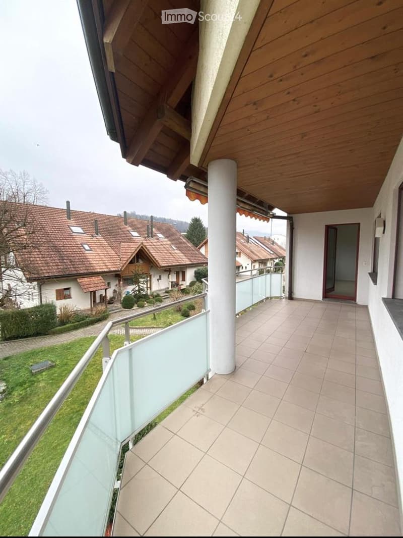 Balcony with tiled floor, wooden ceiling, glass railing, overlooking a residential area with houses and greenery.
