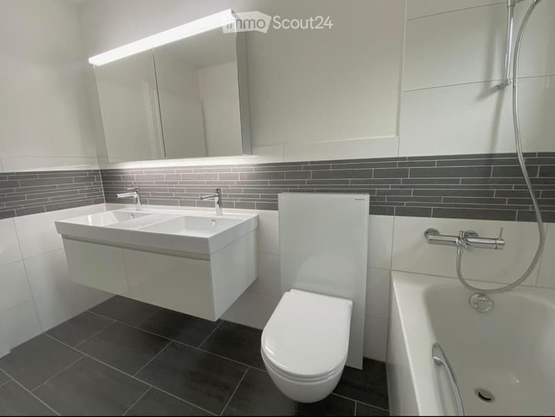 Modern bathroom with a double sink, toilet, bathtub, and large mirror above the sink. Dark tiled floor and walls with a contrasting white vanity and fixtures.