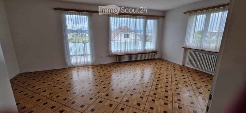 Empty room with wooden parquet flooring, large windows with sheer curtains, radiators, and a view of a house and landscape outside.