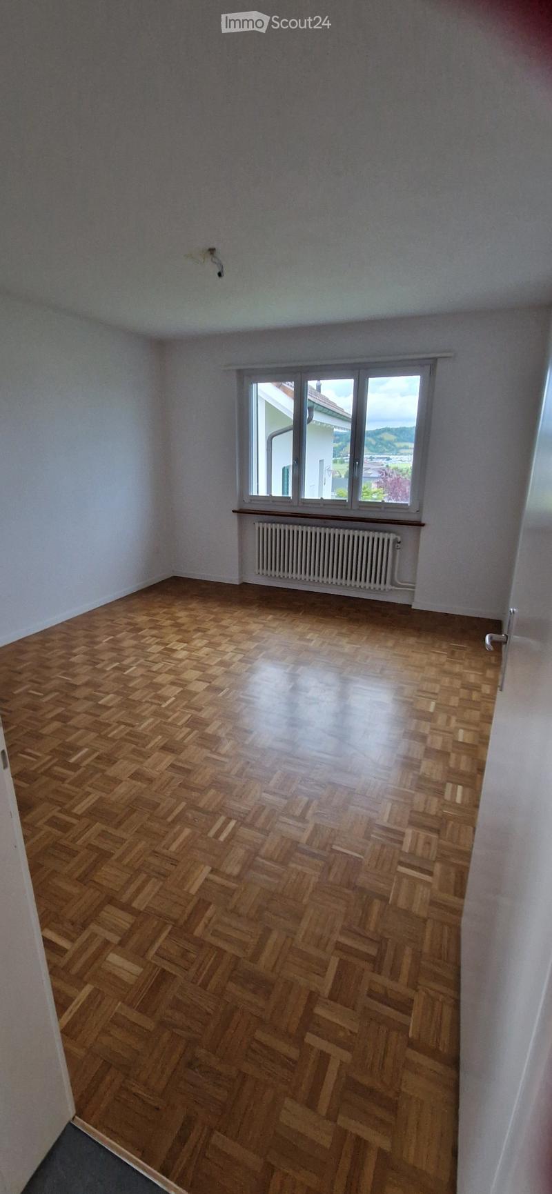 Empty room with wooden herringbone flooring, white walls, and a large window with a radiator below it.