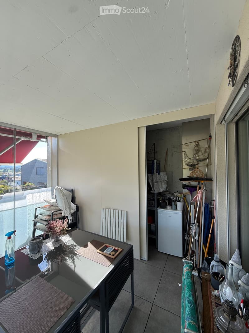 Balcony with a small table, chair, and cleaning supplies stored in a closet. The balcony has a slanted ceiling and a view of the surrounding area.
