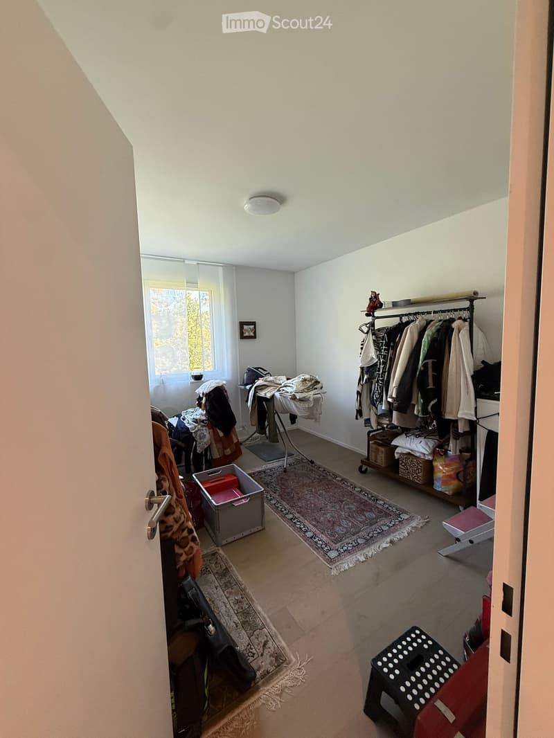 A small, well-lit room with white walls and a slanted ceiling. Contains a clothing rack with various garments, a small stool, a storage box, a table with folded clothes, and rugs on the floor.