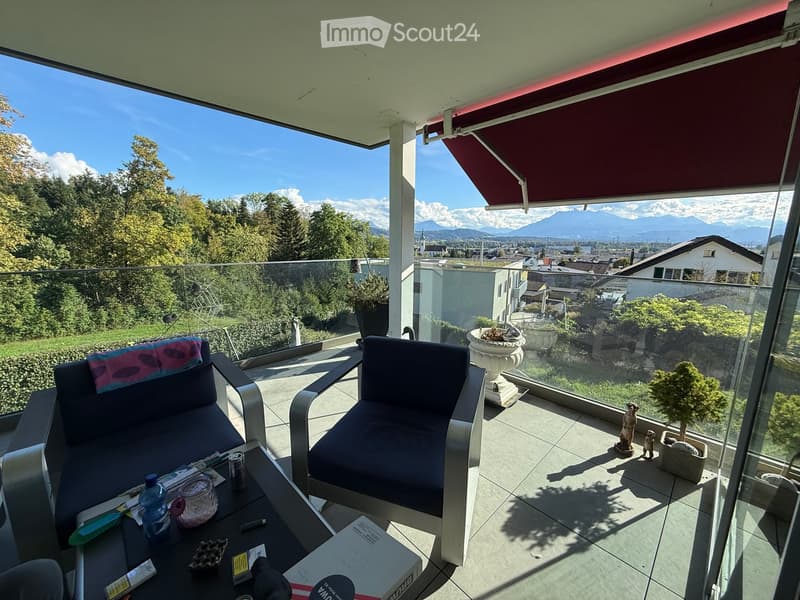 Balcony with a view of mountains and cityscape, featuring a sofa, chair, table, potted plants, and a decorative statue.