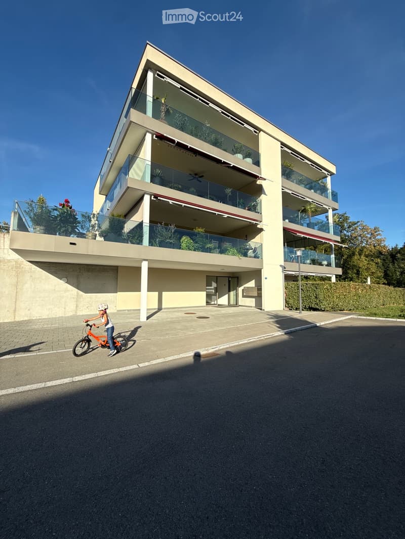 Modern3-story house with large glass balconies, beige facade, and greenery on balconies. A child rides a bicycle in front of the house.