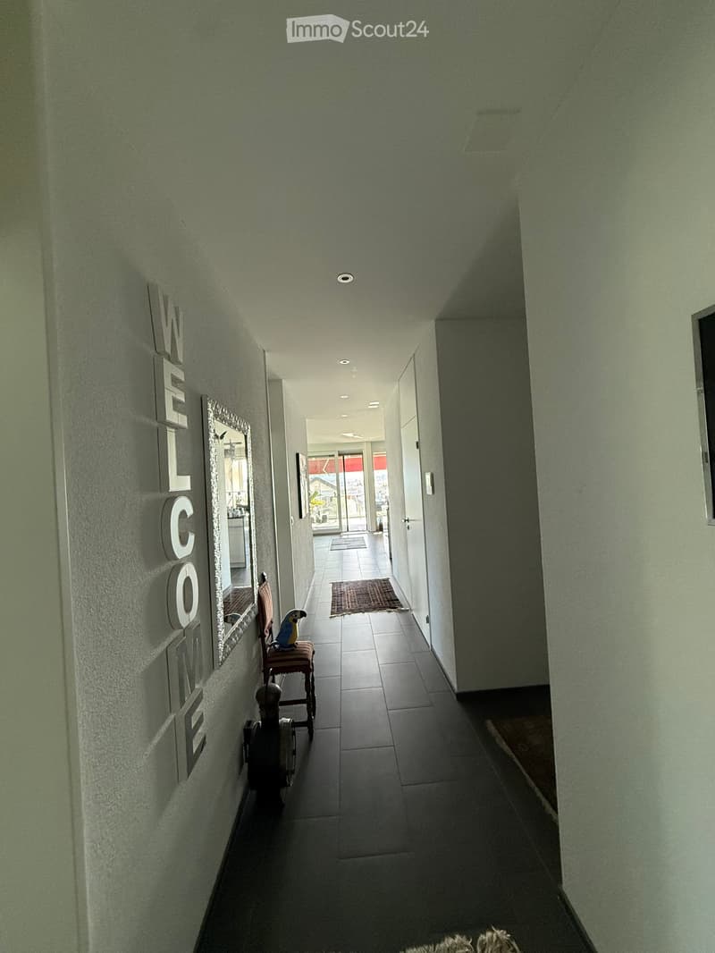 A well-lit hallway with tiled flooring, a small rug, a chair with a parrot, and a mirror on the wall. The word "WELCOME" is displayed on the left wall.