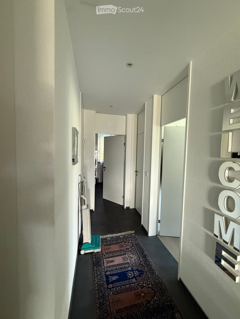 A narrow hallway with light-colored walls, dark flooring, and several doors leading to other rooms. A small rug is placed on the floor, and a decorative sign spelling "WELCOME" is mounted on the right wall.