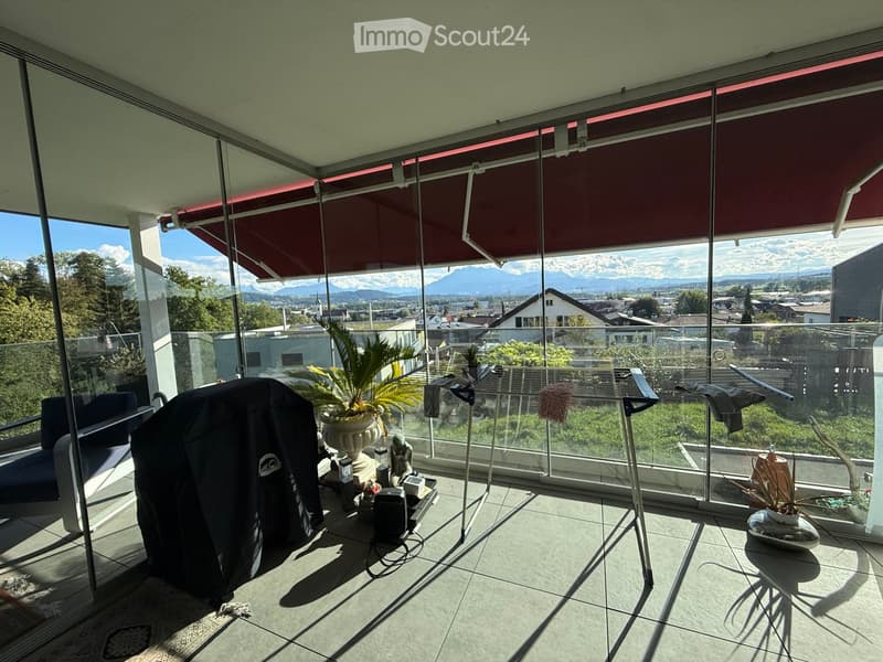 Spacious balcony with large glass walls, offering a scenic mountain view. Features a sofa, small table, potted plants, and a covered grill.