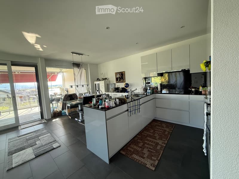 Modern kitchen with white cabinets, island, sink, and various appliances. Features a view through large windows to a balcony area with outdoor furniture.
