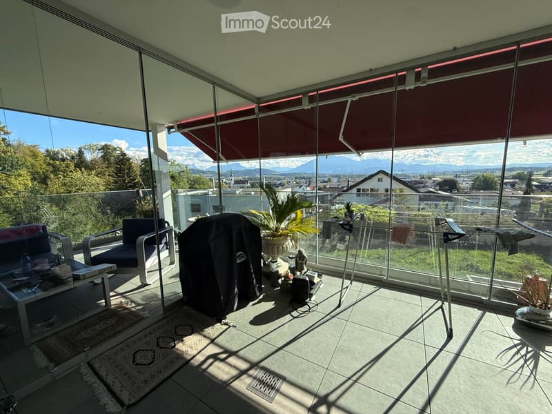 Modern balcony with large glass walls, offering a scenic mountain view, furnished with a sofa, chairs, a small table, and a potted plant.