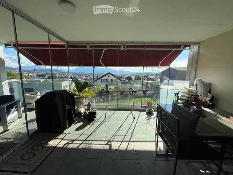 Modern balcony with large glass walls, scenic mountain view, sofa, table, chairs, and plants.