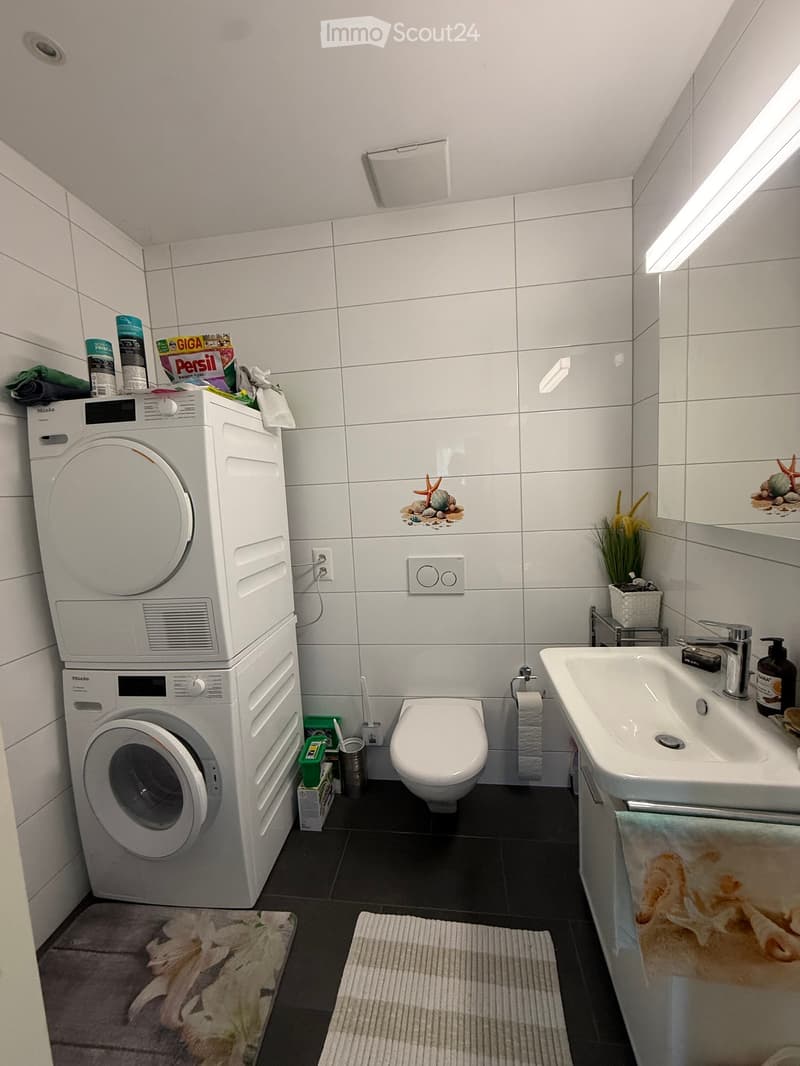 Modern bathroom with white tiles, a stacked washing machine and dryer, toilet, sink, mirror, and decorative items.