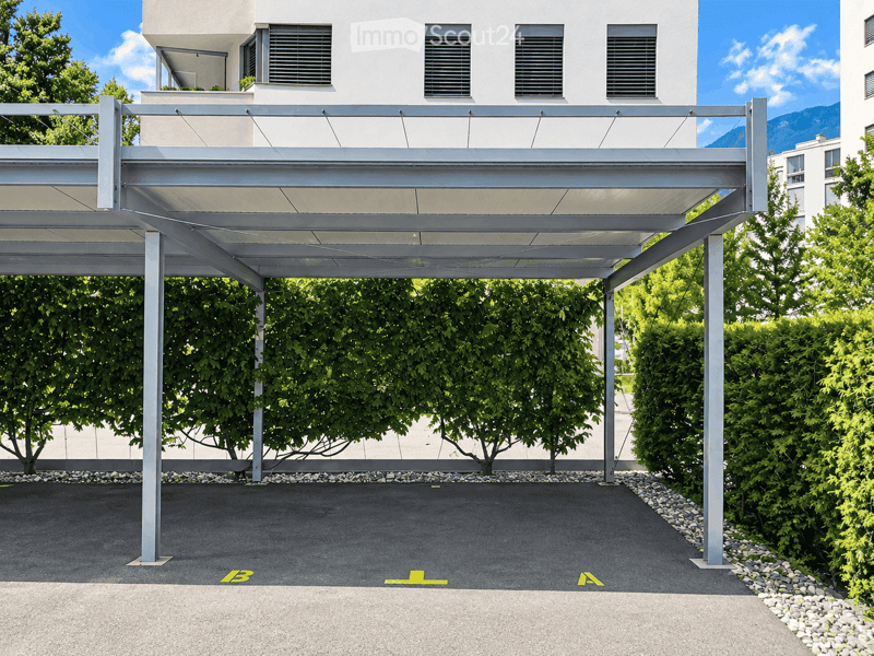 Modern building with a covered parking area, white facade, multiple windows, and greenery surrounding the structure.
