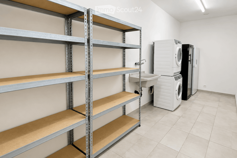 A clean, unfurnished utility room with a washing machine, a sink, a refrigerator, and metal shelving units.