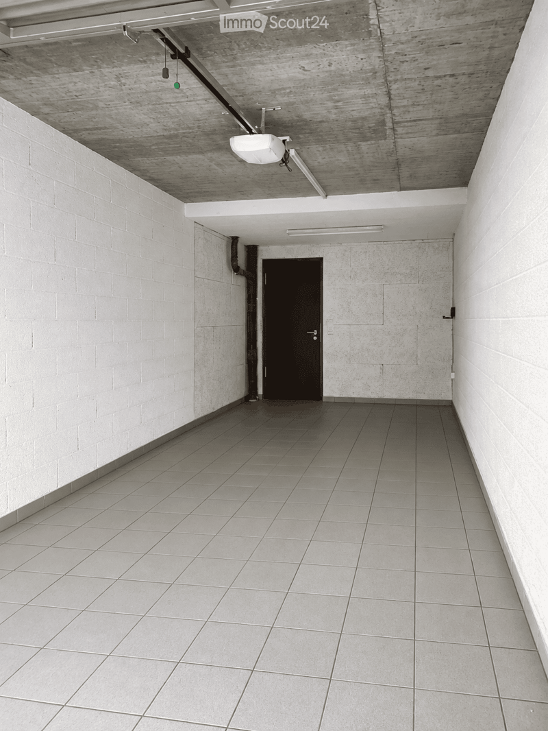 Modern hallway with tiled floor, white brick walls, and a single door at the end. Exposed ceiling beams and integrated lighting fixtures are visible.