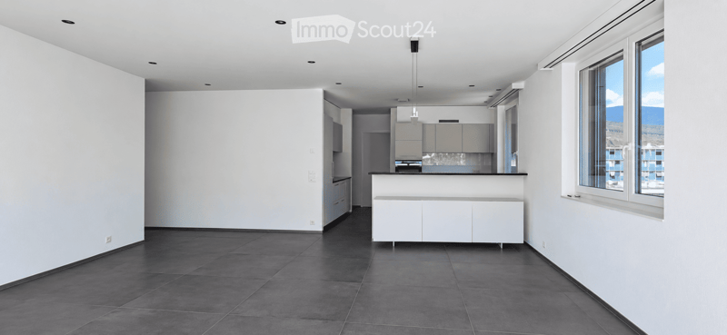 Open-plan unfurnished room with white walls, dark tiled floor, and large windows offering a scenic view. Includes a kitchen area with cabinets and a countertop.