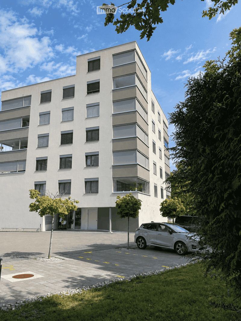 Modern multi-story building with white facade, large windows, and balconies. A car is parked in front, and there are trees and greenery around the building.