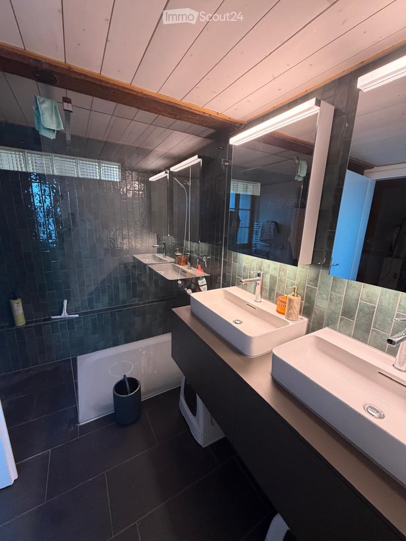 Modern bathroom with two sinks, a large mirror, a shower, and a bathtub. Features dark tiles and wooden beams on the ceiling.