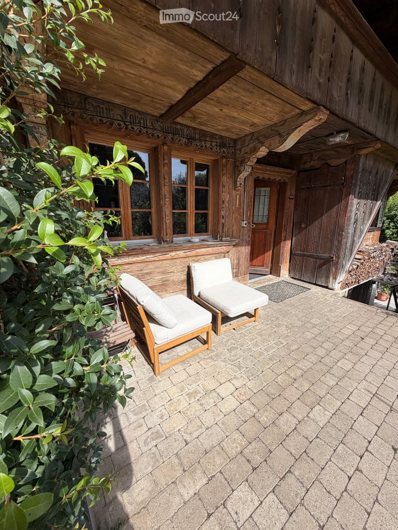A wooden house with a stone-paved patio area featuring two white cushioned chairs and a small table, surrounded by greenery.