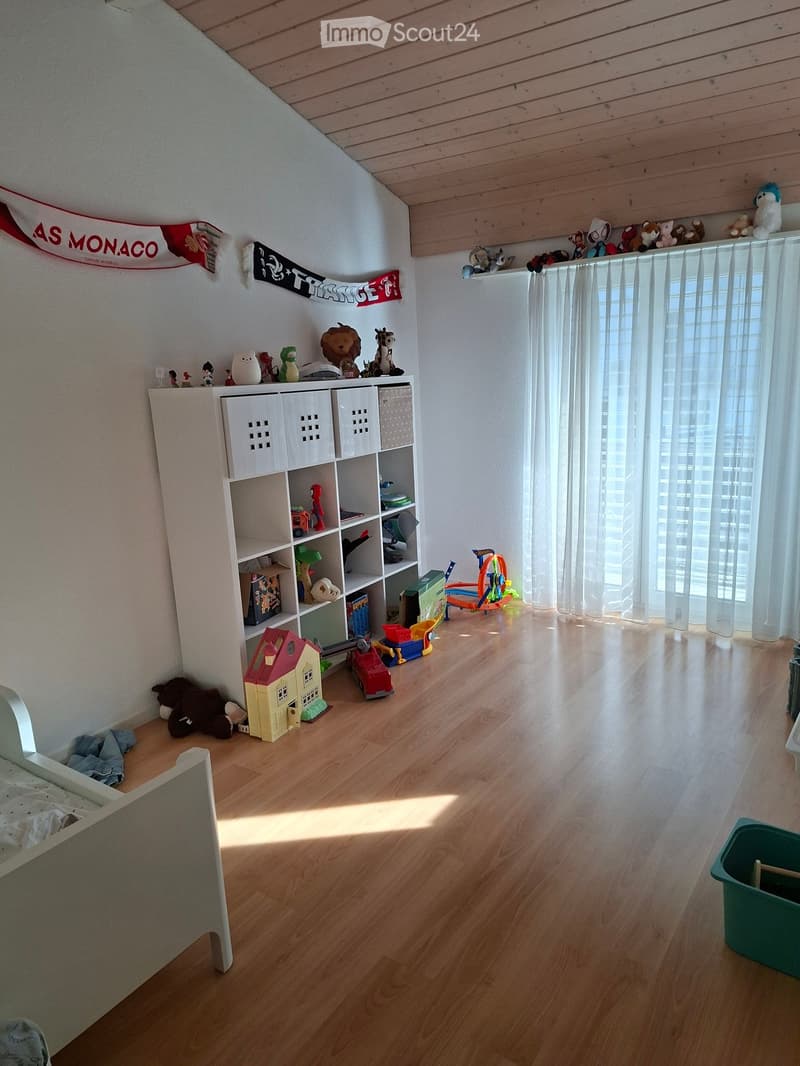 Brightly lit room with wooden flooring, a large window with white curtains, a white shelf filled with toys and books, and a bed visible on the left side.
