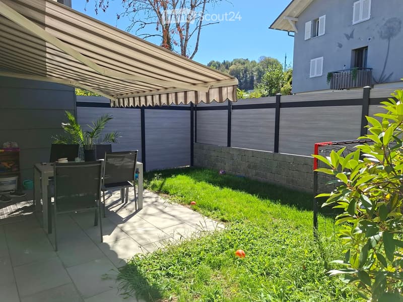 Garden with a covered patio area, a small grassy lawn, a table and chairs, potted plants, and a modern house in the background.