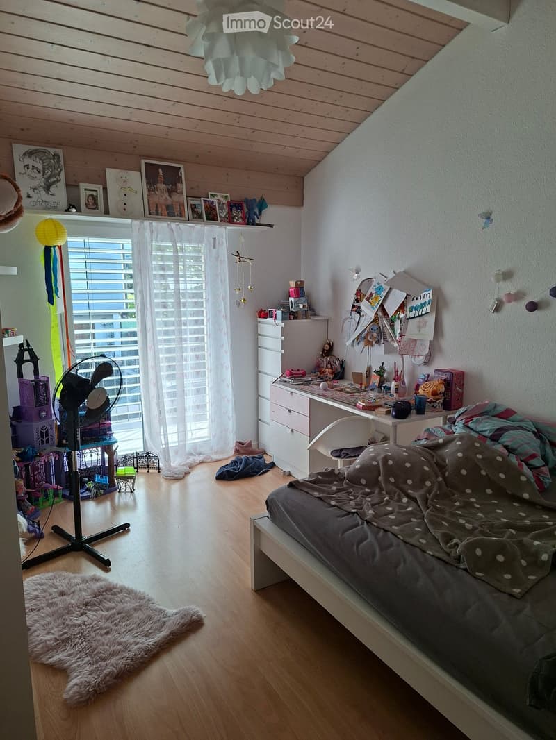 Cozy bedroom with wooden floors, slanted ceiling, bed with grey polka dot blanket, desk with chair, fan, and various toys and decorations.