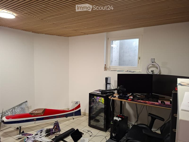 A modern, well-lit room with a desk setup featuring dual monitors, a gaming console, and a gaming chair. A model airplane is displayed on the floor, and a small refrigerator is visible. The room has white walls, a tiled floor, and a window with frosted gl