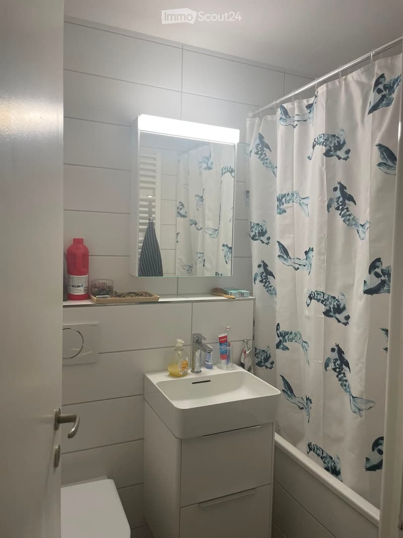 Modern bathroom with white tiles, a sink with a mirror above it, a toilet, and a shower area with a decorative curtain.